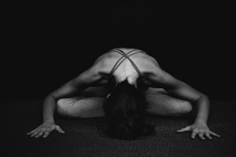 A person in a yoga pose, seated with their legs crossed and bent forward, hands resting on the floor. The setting is minimalistic with a dark background and the focus is on the muscled back and arms.