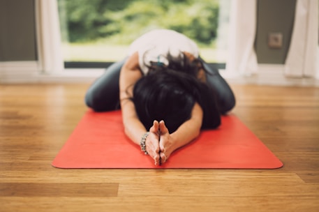 A person is practicing yoga on a red yoga mat indoors. They are in a child's pose position with arms extended forward and forehead touching the mat. The room has a wooden floor and large windows allowing natural light to enter.