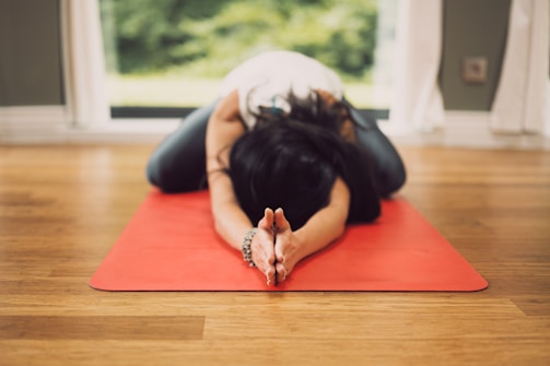 A person is practicing yoga on a red yoga mat indoors. They are in a child's pose position with arms extended forward and forehead touching the mat. The room has a wooden floor and large windows allowing natural light to enter.