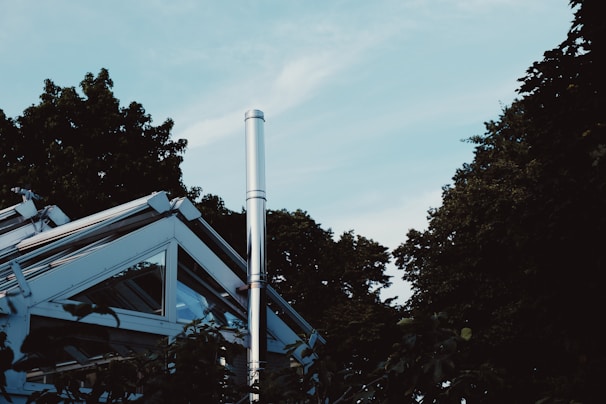 A modern, angular glass and metal structure is surrounded by lush, green trees. The sky above is clear with a pale blue color. A tall, metallic chimney or pipe protrudes from the building, adding an industrial element to the natural setting.
