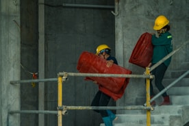 Two construction workers are carrying large red rolls of material up a concrete staircase. Both are wearing safety gear including yellow helmets. The setting appears to be within a building under construction, with concrete walls and metal railings visible.