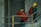 Two construction workers are carrying large red rolls of material up a concrete staircase. Both are wearing safety gear including yellow helmets. The setting appears to be within a building under construction, with concrete walls and metal railings visible.