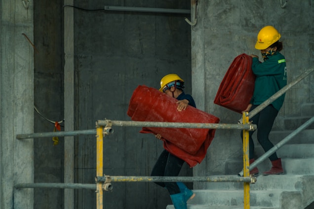 Two construction workers are carrying large red rolls of material up a concrete staircase. Both are wearing safety gear including yellow helmets. The setting appears to be within a building under construction, with concrete walls and metal railings visible.