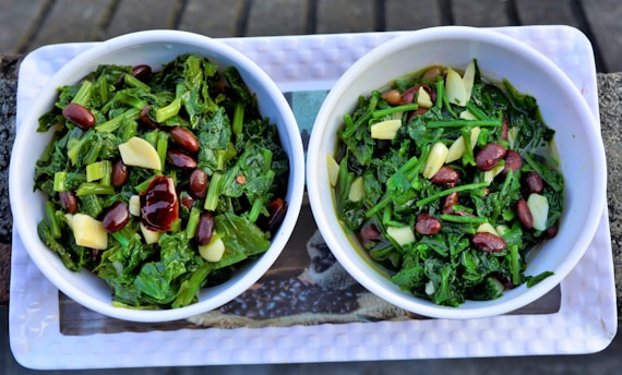 Two bowls filled with a green leafy salad composed of chopped greens, red kidney beans, and slices of garlic. The bowls are white and placed on a rectangular tray.