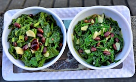 Two bowls filled with a green leafy salad composed of chopped greens, red kidney beans, and slices of garlic. The bowls are white and placed on a rectangular tray.