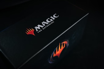 A black box featuring the bold logo and text 'Magic: The Gathering' in white, accompanied by a striking red and orange flame-like emblem.