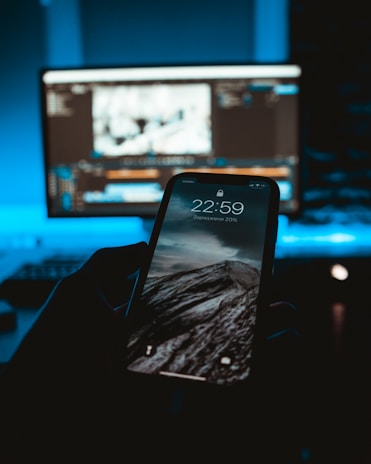 A smartphone displaying a mountain landscape on its screen is held in a dark room. In the background, a computer monitor shows a blurred image or video editing software, casting a blue glow.