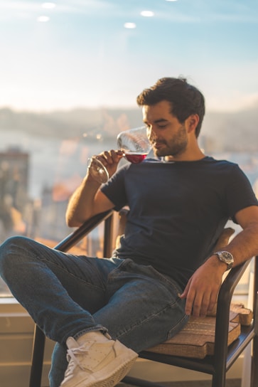 A man is sitting in a chair, holding and smelling a glass of red wine. He is wearing a dark blue t-shirt, blue jeans, white sneakers, and a watch. The background shows a distant and blurred urban landscape seen through a large window, with soft, warm lighting creating a relaxed atmosphere.