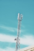 A telecommunications tower extends into a clear blue sky with minimal clouds. The structure features metal beams and antennae, indicating its use for broadcasting or communication purposes.