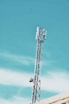 A telecommunications tower extends into a clear blue sky with minimal clouds. The structure features metal beams and antennae, indicating its use for broadcasting or communication purposes.