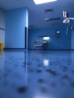 A hospital room with blue walls and a reflective floor. An empty hospital bed is positioned against the wall, surrounded by medical equipment and monitors. The lighting is bright, creating a clean and sterile environment.