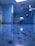 A hospital room with blue walls and a reflective floor. An empty hospital bed is positioned against the wall, surrounded by medical equipment and monitors. The lighting is bright, creating a clean and sterile environment.