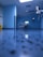 A hospital room with blue walls and a reflective floor. An empty hospital bed is positioned against the wall, surrounded by medical equipment and monitors. The lighting is bright, creating a clean and sterile environment.