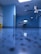 A hospital room with blue walls and a reflective floor. An empty hospital bed is positioned against the wall, surrounded by medical equipment and monitors. The lighting is bright, creating a clean and sterile environment.