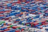 Aerial view of a large shipping yard filled with numerous colorful cargo containers arranged in neat rows. The containers display various company names and logos, with colors including blue, red, green, and yellow, creating a vivid patchwork. The scene is busy yet organized, indicative of a hub for goods transportation.