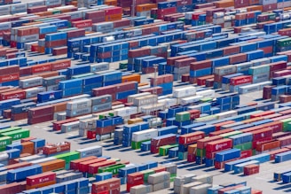 Aerial view of a large shipping yard filled with numerous colorful cargo containers arranged in neat rows. The containers display various company names and logos, with colors including blue, red, green, and yellow, creating a vivid patchwork. The scene is busy yet organized, indicative of a hub for goods transportation.