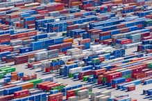 Aerial view of a large shipping yard filled with numerous colorful cargo containers arranged in neat rows. The containers display various company names and logos, with colors including blue, red, green, and yellow, creating a vivid patchwork. The scene is busy yet organized, indicative of a hub for goods transportation.