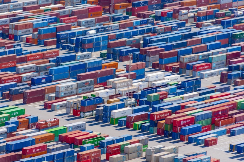 Aerial view of a large shipping yard filled with numerous colorful cargo containers arranged in neat rows. The containers display various company names and logos, with colors including blue, red, green, and yellow, creating a vivid patchwork. The scene is busy yet organized, indicative of a hub for goods transportation.