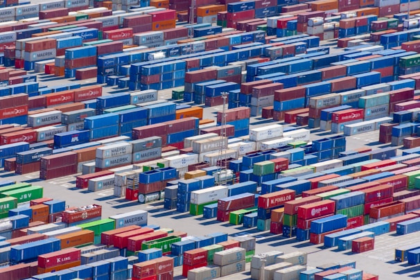 Aerial view of a large shipping yard filled with numerous colorful cargo containers arranged in neat rows. The containers display various company names and logos, with colors including blue, red, green, and yellow, creating a vivid patchwork. The scene is busy yet organized, indicative of a hub for goods transportation.