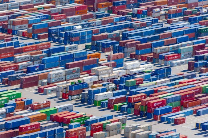 Aerial view of a large shipping yard filled with numerous colorful cargo containers arranged in neat rows. The containers display various company names and logos, with colors including blue, red, green, and yellow, creating a vivid patchwork. The scene is busy yet organized, indicative of a hub for goods transportation.