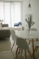 A minimalist living room with a modern design features a round white dining table surrounded by white chairs with wooden legs. A clear glass vase with eucalyptus branches sits on the table. In the background, a gray sofa is placed against the wall near a large window with horizontal blinds, allowing soft light to filter in. A blue blanket drapes over a white armchair, and a hanging pendant light adds a touch of industrial style.