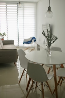A minimalist living room with a modern design features a round white dining table surrounded by white chairs with wooden legs. A clear glass vase with eucalyptus branches sits on the table. In the background, a gray sofa is placed against the wall near a large window with horizontal blinds, allowing soft light to filter in. A blue blanket drapes over a white armchair, and a hanging pendant light adds a touch of industrial style.