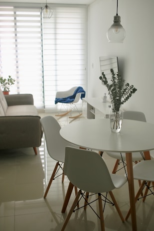 A minimalist living room with a modern design features a round white dining table surrounded by white chairs with wooden legs. A clear glass vase with eucalyptus branches sits on the table. In the background, a gray sofa is placed against the wall near a large window with horizontal blinds, allowing soft light to filter in. A blue blanket drapes over a white armchair, and a hanging pendant light adds a touch of industrial style.