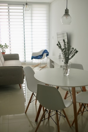 A minimalist living room with a modern design features a round white dining table surrounded by white chairs with wooden legs. A clear glass vase with eucalyptus branches sits on the table. In the background, a gray sofa is placed against the wall near a large window with horizontal blinds, allowing soft light to filter in. A blue blanket drapes over a white armchair, and a hanging pendant light adds a touch of industrial style.