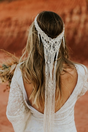 A woman with long hair is adorned with a detailed macrame hair accessory, showcasing intricate patterns. She is wearing an elegant, lace-textured white dress with an open back. In her hand, she holds a bouquet of dried flowers. The background features a warm, earthy, reddish-brown landscape, suggesting an outdoor setting, possibly a desert or canyon.