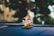 A small statue of Ganesha with a golden headdress and colorful garments sits on a car dashboard. The background is blurred, showing hints of greenery and sunlight.