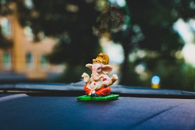 A small statue of Ganesha with a golden headdress and colorful garments sits on a car dashboard. The background is blurred, showing hints of greenery and sunlight.