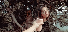 A hand holding a round mirror reflects a woman's face in a lush, leafy outdoor setting. The woman's eyes are closed, her lips slightly parted, with an expression of serenity.