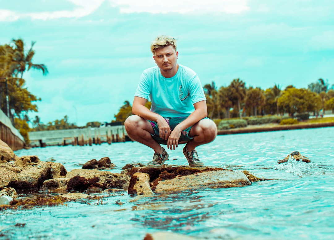 man in teal crew neck t-shirt sitting on brown rock in water during daytime