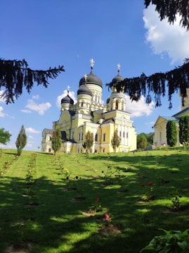 A beautiful Orthodox church with multiple domes, topped by crosses, stands prominently in the background. The building features light yellow walls with arched windows, surrounded by well-manicured gardens. The foreground shows a lush green lawn with rows of small, colorful flowers. The sky is clear with a few cumulus clouds, and pine tree branches frame the image.