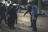 A group of elderly people are gathered in a park, playing a game of pétanque. They are focused on the arrangement of metal balls scattered on the sandy ground. One person is pointing towards the balls as others observe intently, demonstrating the social and engaging nature of the game.