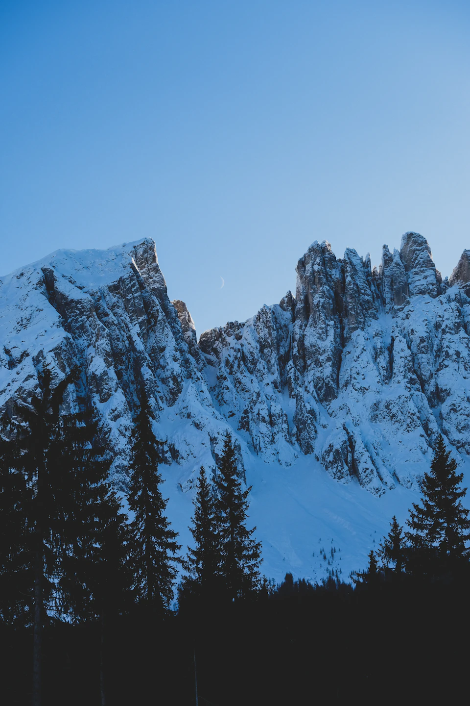 The image depicts a majestic mountain range under a clear blue sky, with snow-capped peaks reaching towards the heavens. The mountains are blanketed in a thick layer of snow and stand tall against the backdrop of the sky. In the foreground, there is a dense forest filled with trees that add depth to the scene. The perspective of the image is from below, looking up at the towering mountains, creating a sense of awe and grandeur.