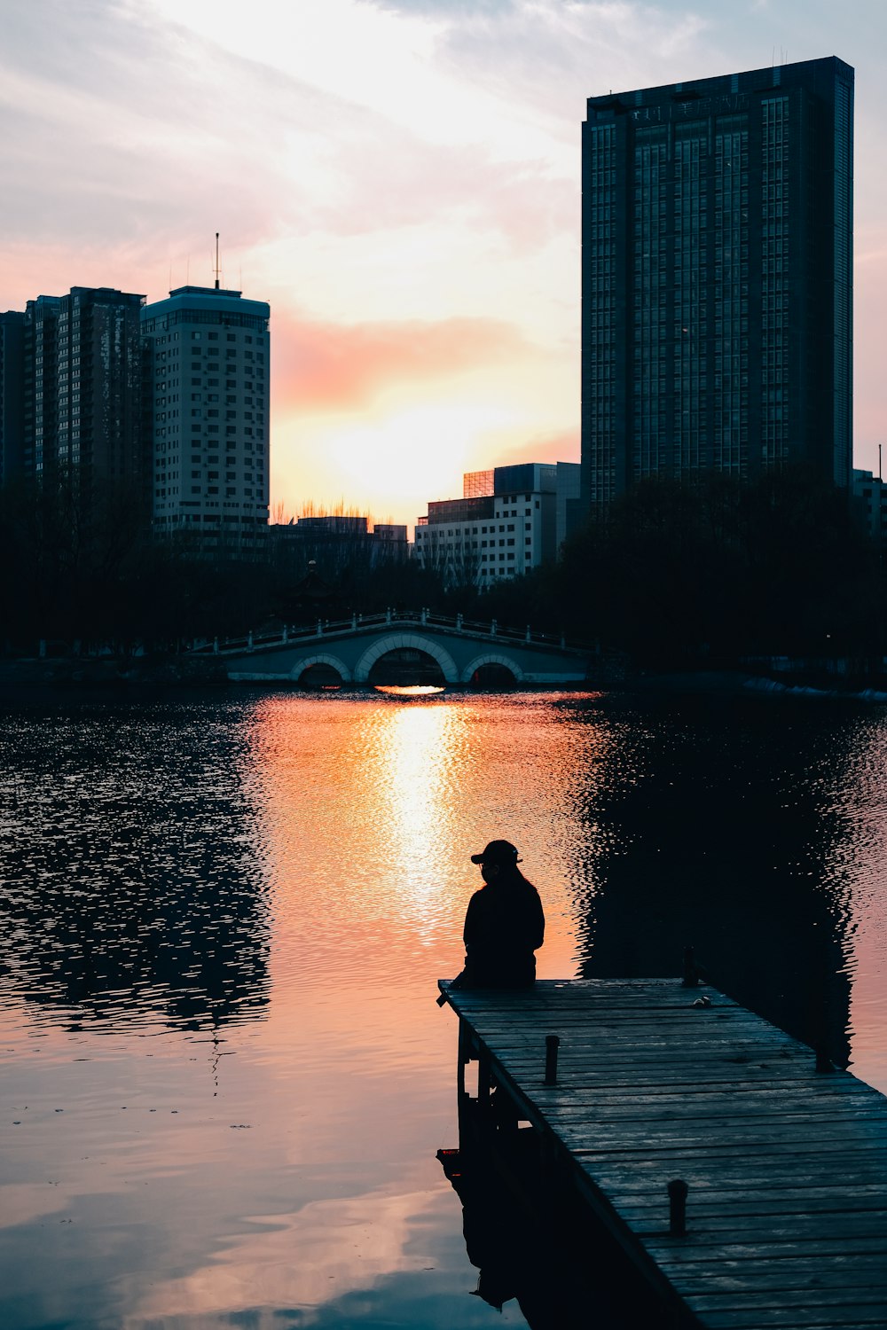 man sitting on wooden dock near body of water during sunset