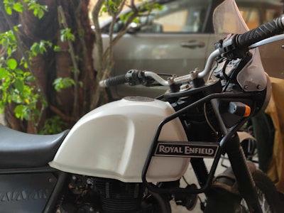 A motorcycle with a white fuel tank and black frame, prominently displaying the Royal Enfield logo. The bike is parked near a tree with vibrant green foliage and a car in the background. The handlebar grips and dashboard are visible, indicating it is a Royal Enfield Himalayan model.