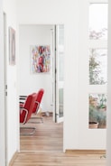 A bright, modern interior with white walls and wooden flooring. Red chairs with metal legs are aligned in the room. There is a colorful abstract painting on the wall and a few potted plants are visible through a large window, adding greenery to the space.