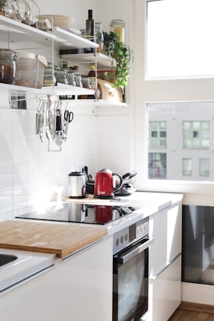 A bright, modern kitchen with white cabinets and an electric stove. Sunlight streams through a large window, illuminating the countertop which features a wooden cutting board and a red electric kettle. Open shelving displays neatly organized jars, bottles, and a potted plant, along with hanging kitchen utensils.