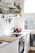 A bright, modern kitchen with white cabinets and an electric stove. Sunlight streams through a large window, illuminating the countertop which features a wooden cutting board and a red electric kettle. Open shelving displays neatly organized jars, bottles, and a potted plant, along with hanging kitchen utensils.