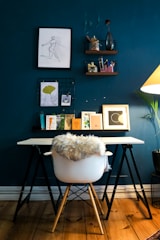 A stylish workspace featuring a white chair with a fur throw draped over it, positioned at a minimalist desk. The desk is supported by black trestles and displays a collection of framed art, books, and decorative items. The dark teal wall behind hosts a sketch in a black frame, along with hanging shelves that hold stationery and small plants. A warm lamp and a hint of greenery add to the ambience.