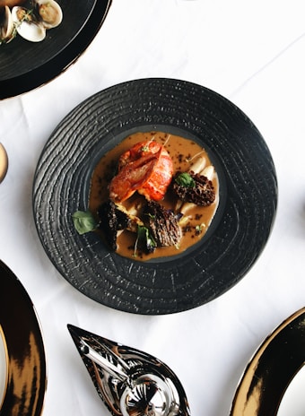A beautifully plated gourmet dish with what appears to be a piece of lobster, morel mushrooms, and edible greens, served on a black textured plate. The plate is set on a white tablecloth with other black dishes and silverware visible around it.