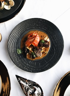 A beautifully plated gourmet dish with what appears to be a piece of lobster, morel mushrooms, and edible greens, served on a black textured plate. The plate is set on a white tablecloth with other black dishes and silverware visible around it.