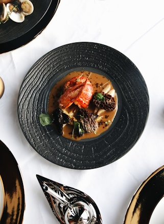 A beautifully plated gourmet dish with what appears to be a piece of lobster, morel mushrooms, and edible greens, served on a black textured plate. The plate is set on a white tablecloth with other black dishes and silverware visible around it.