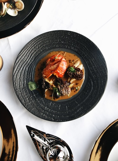 A beautifully plated gourmet dish with what appears to be a piece of lobster, morel mushrooms, and edible greens, served on a black textured plate. The plate is set on a white tablecloth with other black dishes and silverware visible around it.