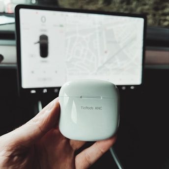 A hand holds a white TicPods ANC case in the foreground. The background features a vehicle's dashboard with a digital display showing a map interface.