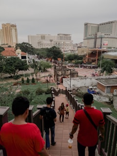 A group of people walking down a staircase towards a historic gate located in a city setting. Tall modern buildings and lush green trees surround the area. The scene is bustling with activity as other people can be seen in the background near the historic structure.