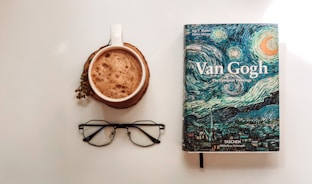 A visually appealing arrangement featuring a mug of frothy coffee resting on a coaster, positioned near a book titled 'Van Gogh: The Complete Paintings.' The book cover displays a vibrant depiction inspired by Van Gogh's style, including swirling night skies. Nearby, a pair of black-rimmed glasses adds a sense of intellectual ambiance.