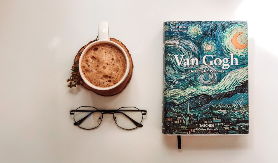 A visually appealing arrangement featuring a mug of frothy coffee resting on a coaster, positioned near a book titled 'Van Gogh: The Complete Paintings.' The book cover displays a vibrant depiction inspired by Van Gogh's style, including swirling night skies. Nearby, a pair of black-rimmed glasses adds a sense of intellectual ambiance.